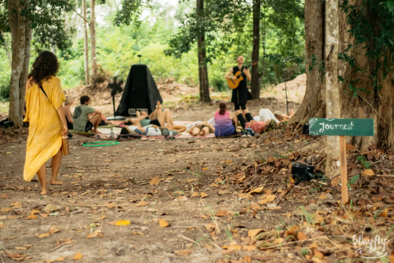 A guitarist plays for listeners resting on mats beneath the trees, a Journest sign in the foreground.