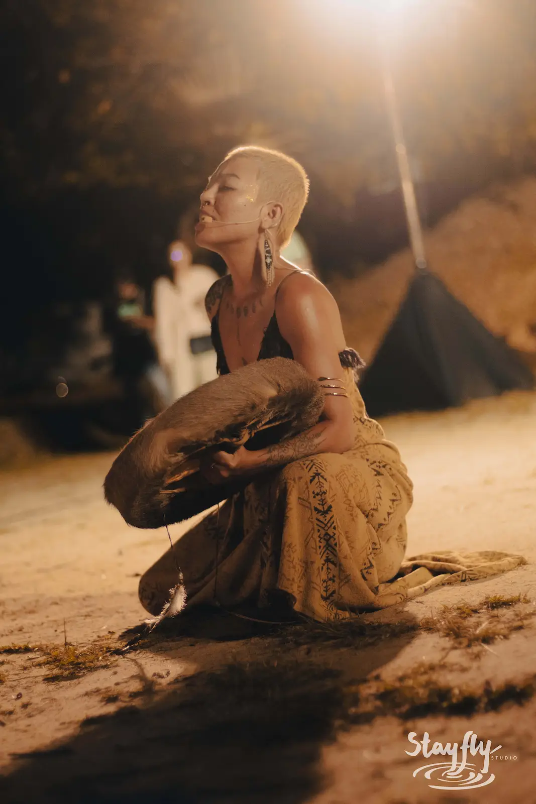 Bald woman kneeling with drum at night ceremony.