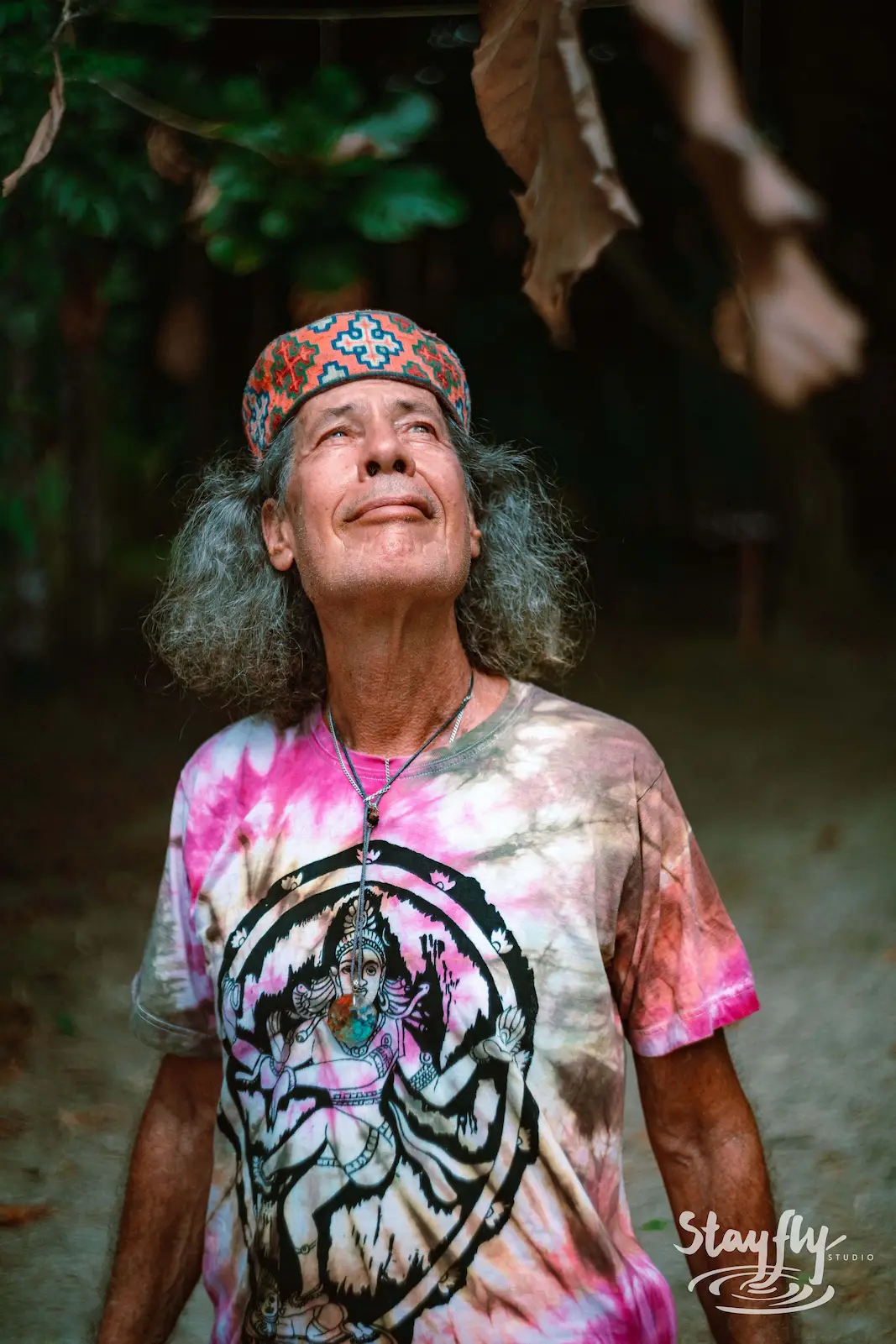 Older man in tie-dye shirt looking up in forest.