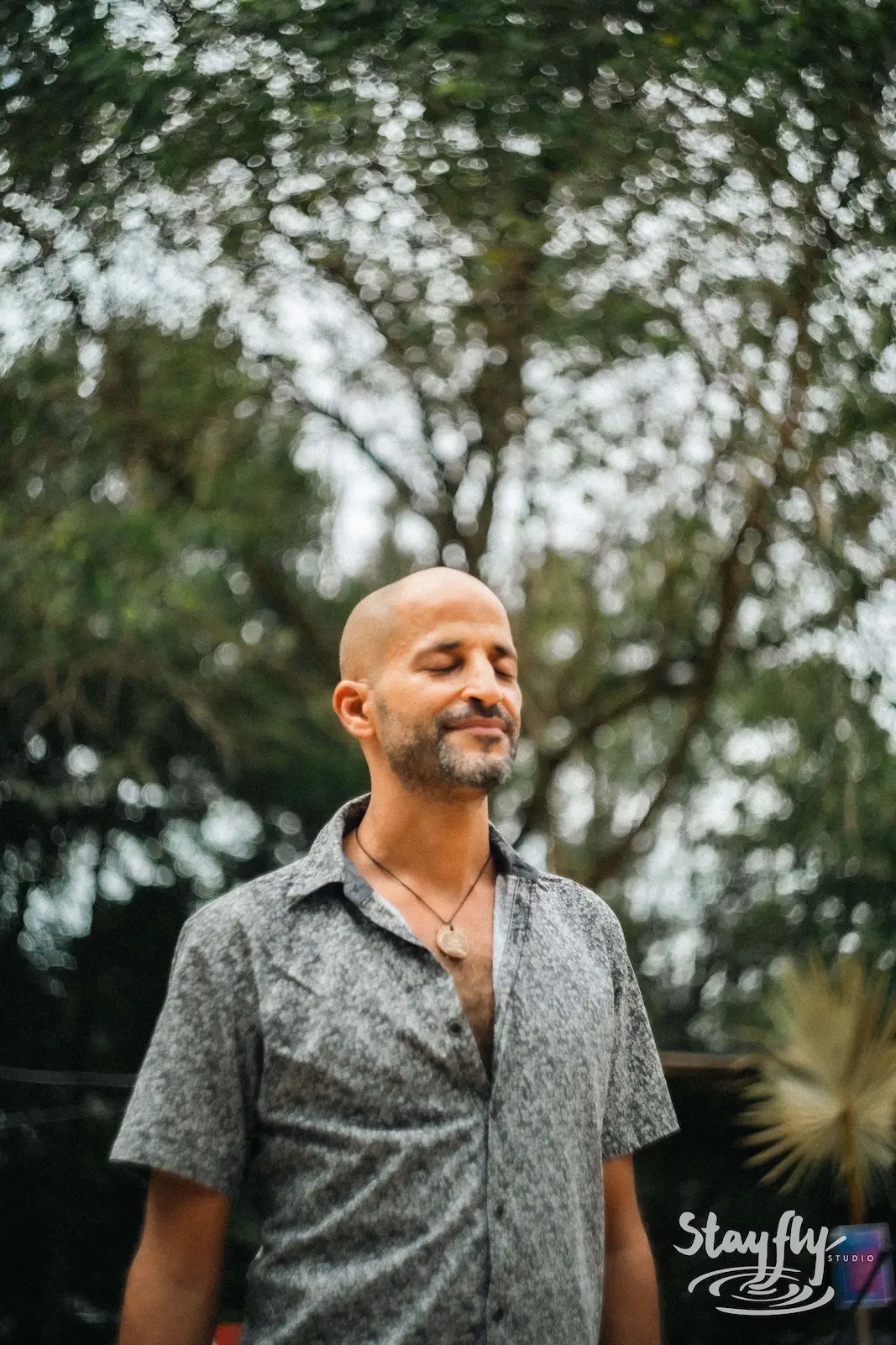Man with shaved head smiling among trees.