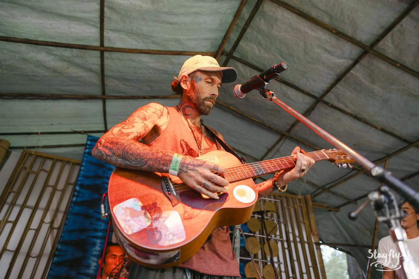 Tattooed guitarist singing under tent roof.