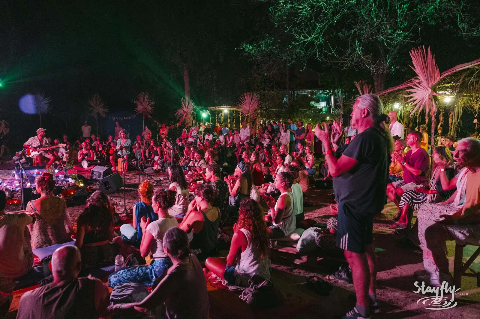 Man gestures to crowd under red stage lighting.
