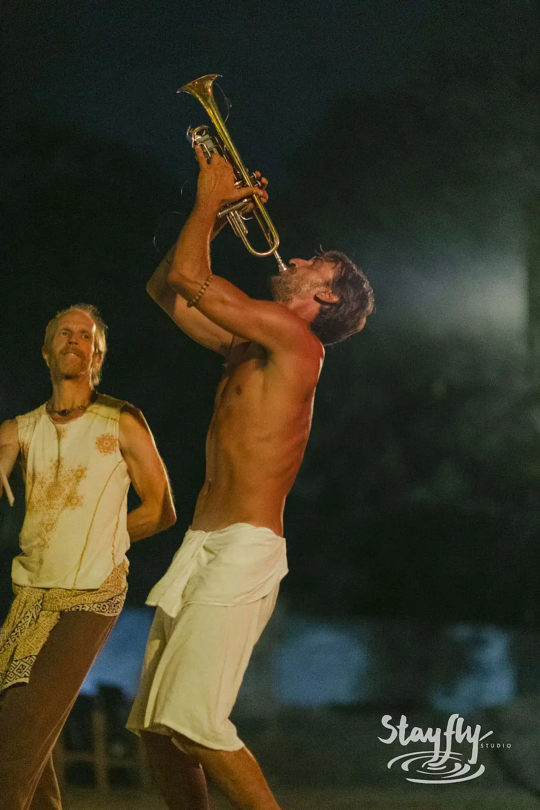Shirtless man playing trumpet skyward at night.