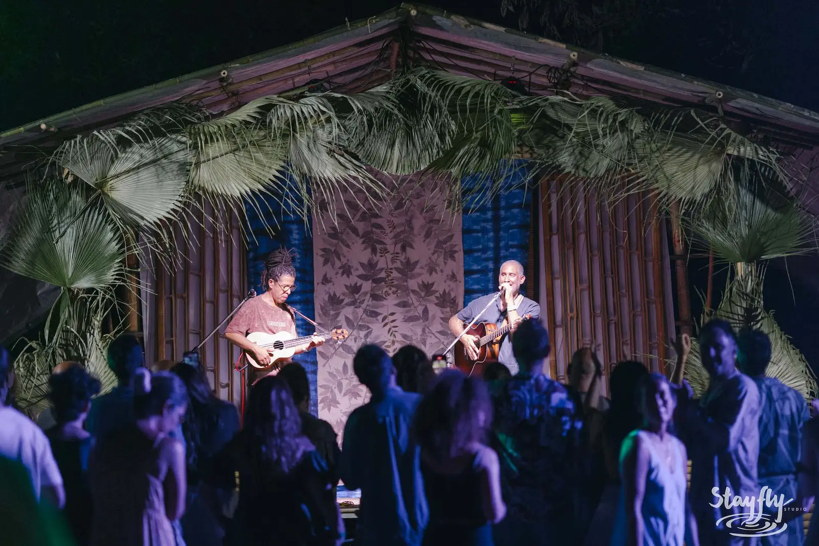 Two guitarists on palm-thatched stage facing crowd.