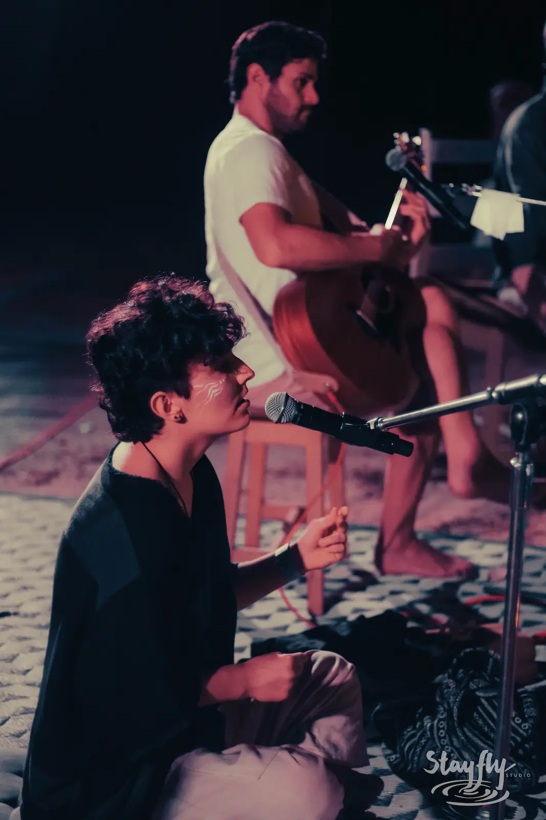 Singer kneeling at mic with guitarist behind at night.