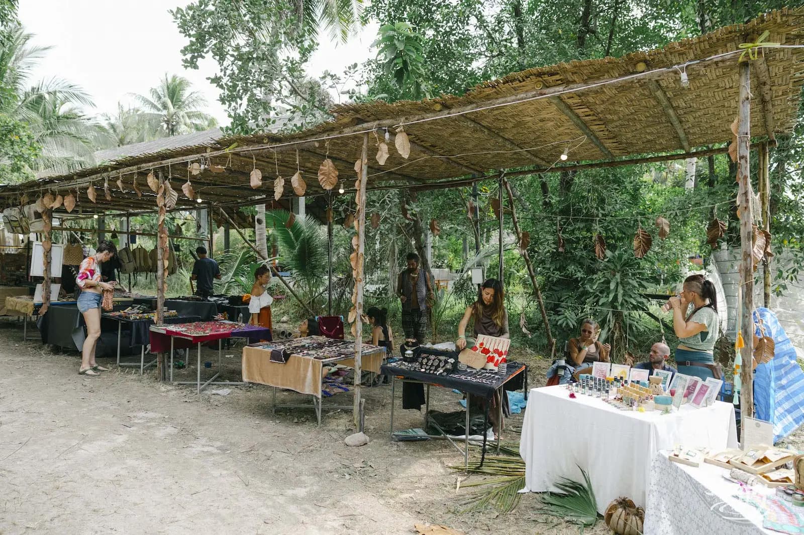 Row of market stalls under thatched roof by trees.