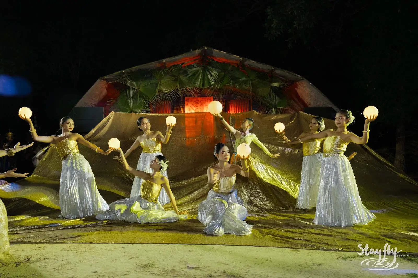 Dancers in white and gold holding glowing orbs at night.
