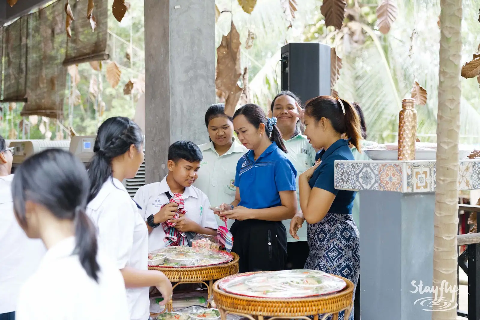 Students and teachers gather around woven trays of food at the school's stall, leaves hanging above them.