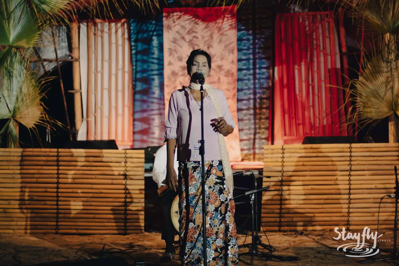 Woman speaking at mic on decorated bamboo stage.