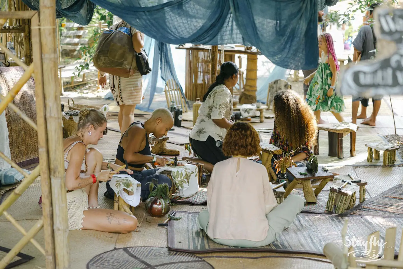 People crafting seated on mats under blue tarp.