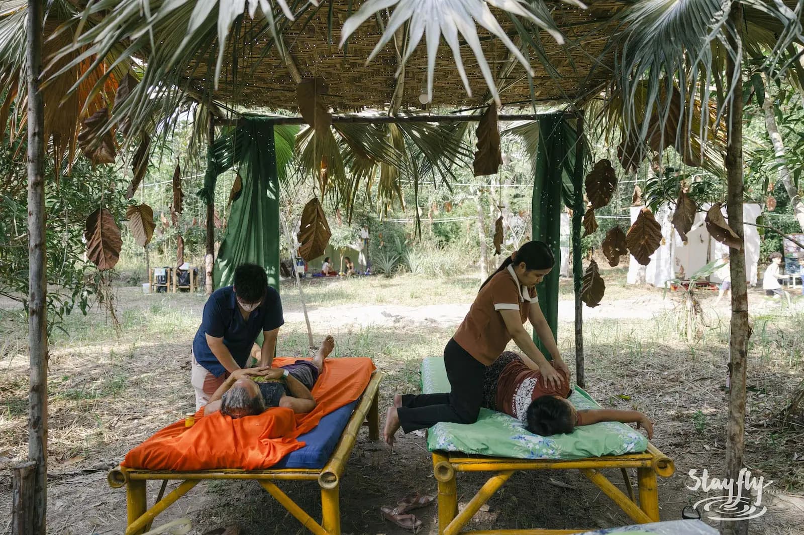 Two practitioners work with participants on treatment tables beneath a palm-leaf shelter.