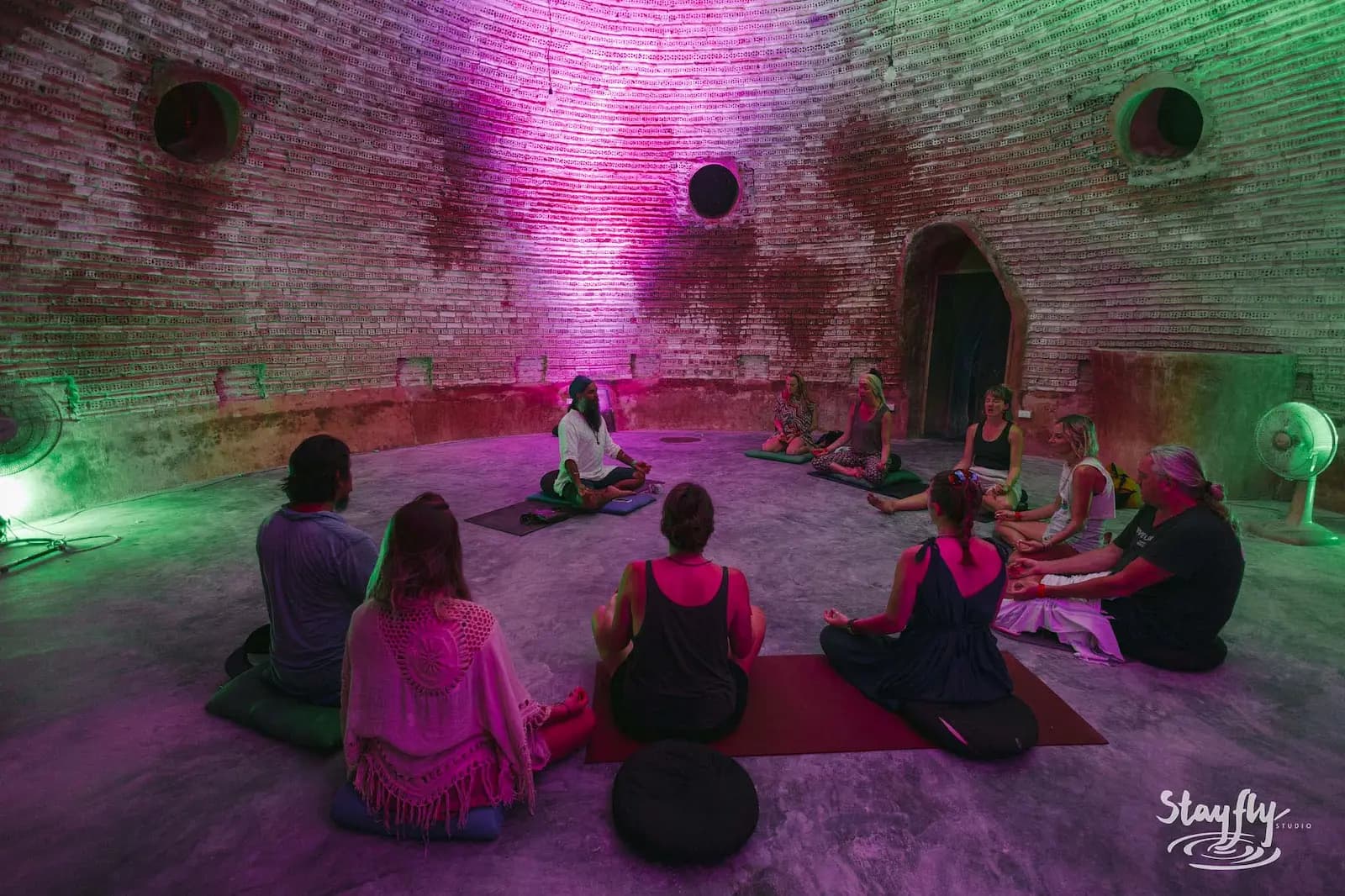 People seated on mats inside a round brick room lit pink and green.