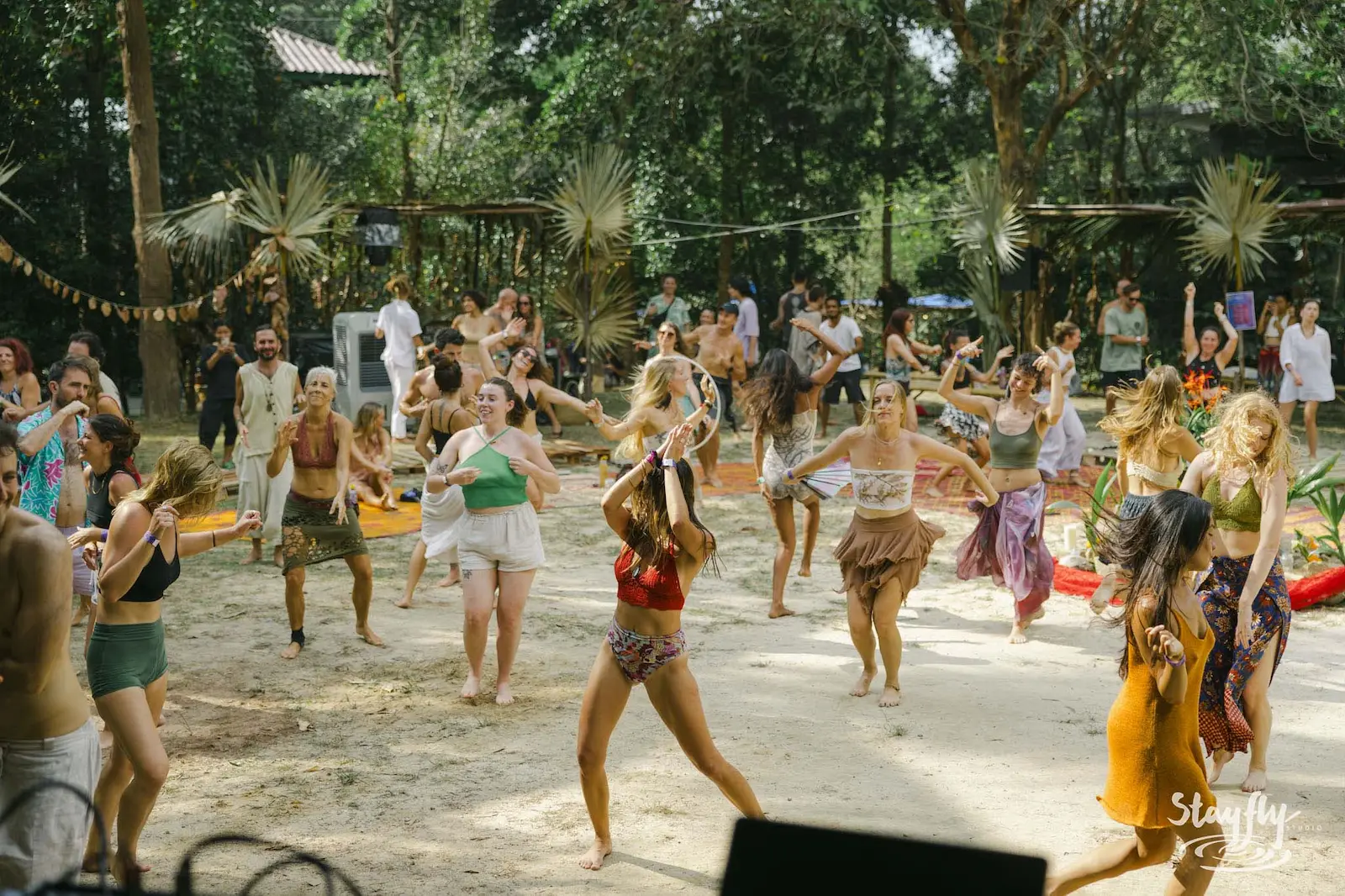 People dancing together on sandy ground among trees in daylight.