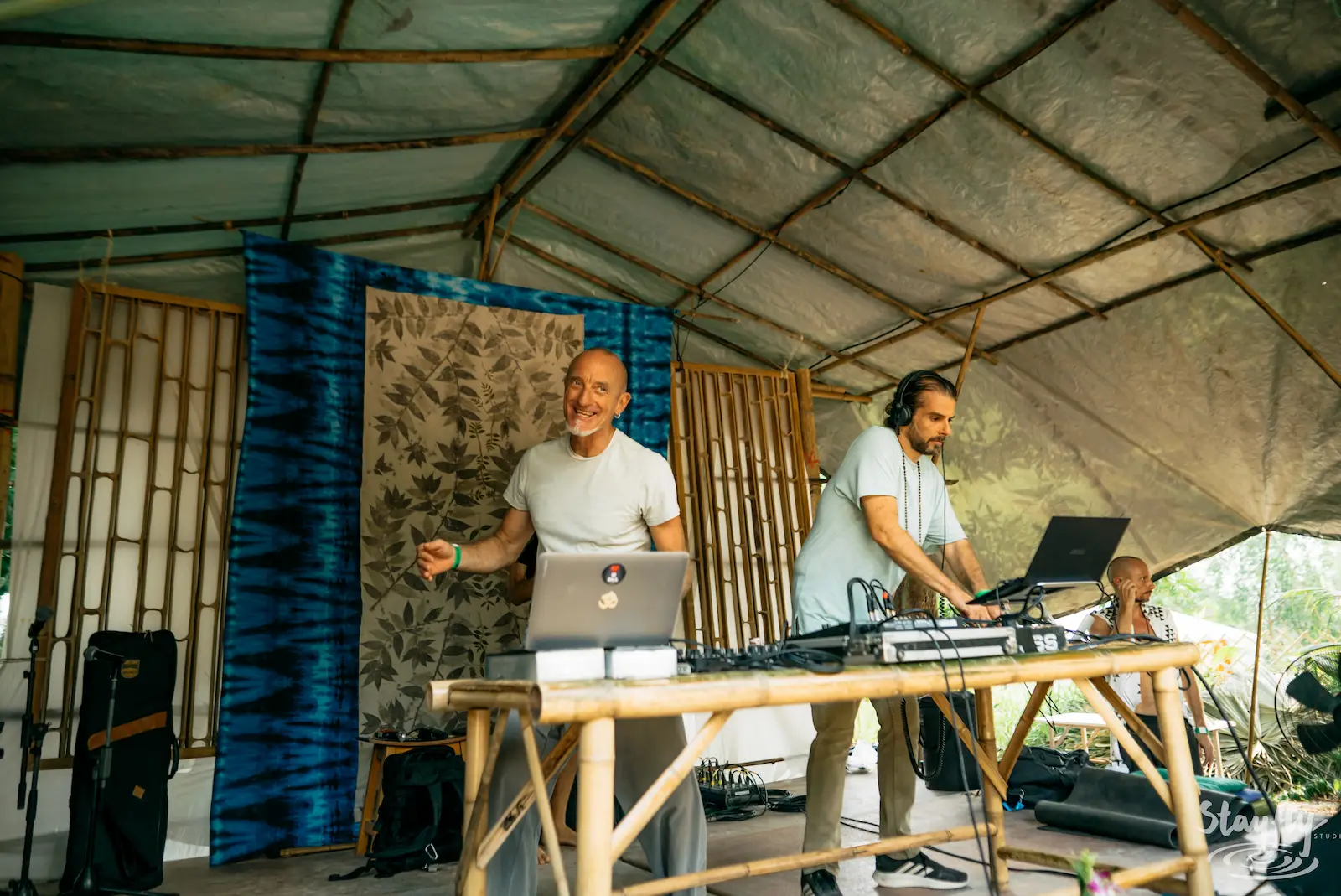 Two DJs stand behind laptops and audio equipment on a covered bamboo stage.