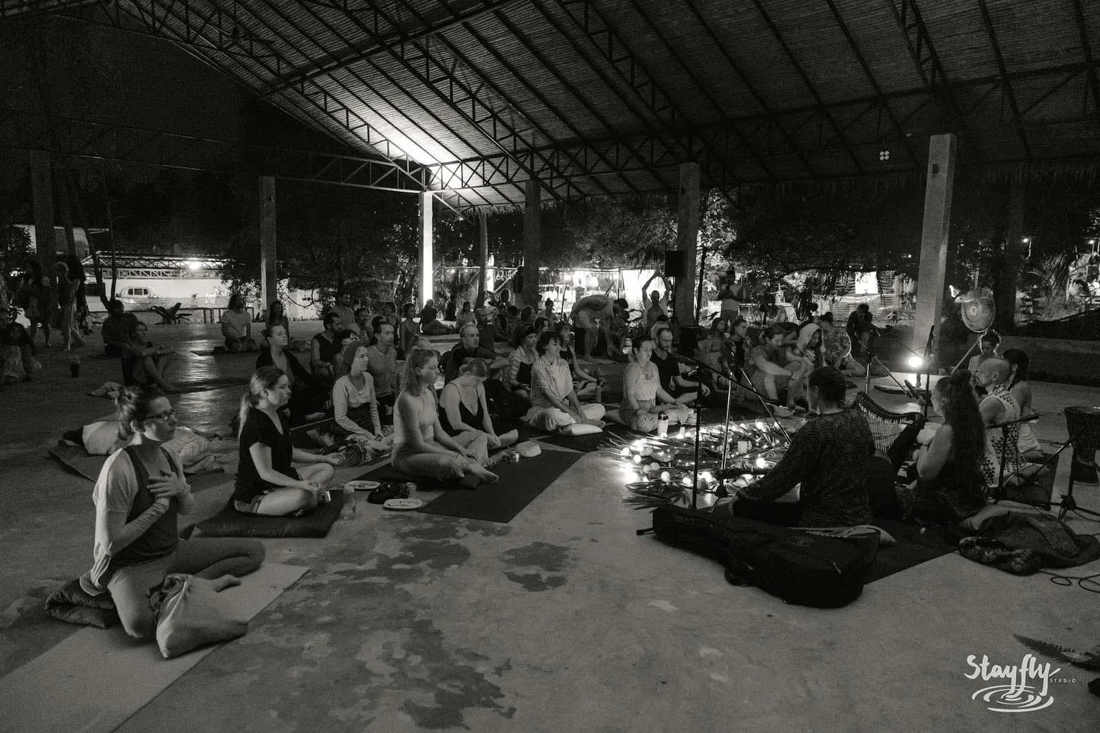 Black-and-white photograph of seated musicians and an audience in a large open-sided hall at night.