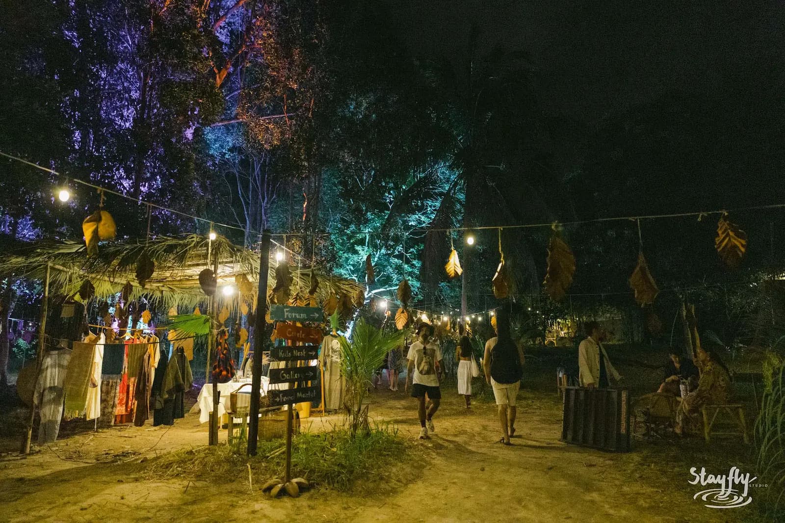 People walk past lit stalls, leaf decorations and a colourful direction sign at night.