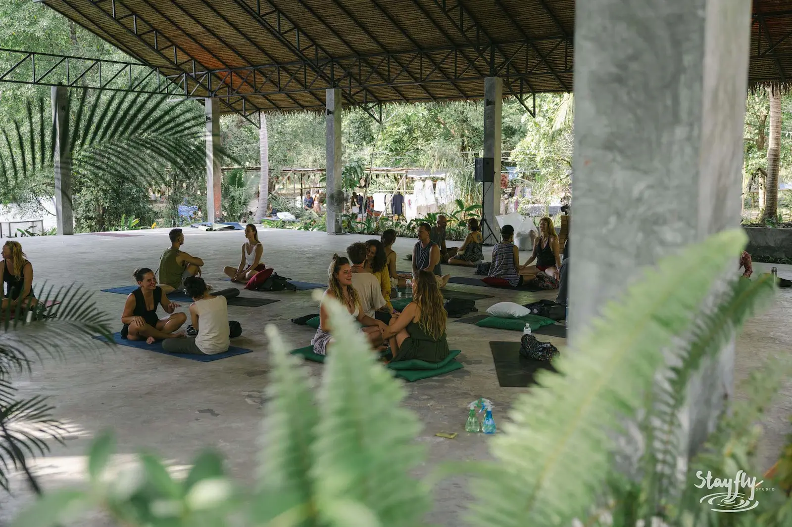 Small groups sit on mats in an open-sided hall, seen past green leaves.