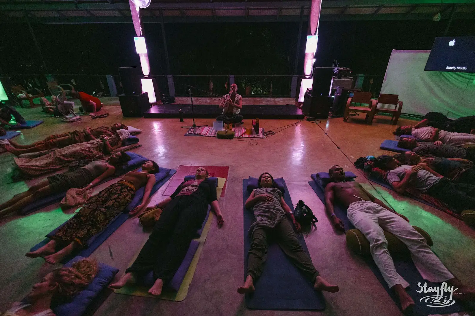 Participants lie on mats around a seated sound performer in a hall lit pink and green.