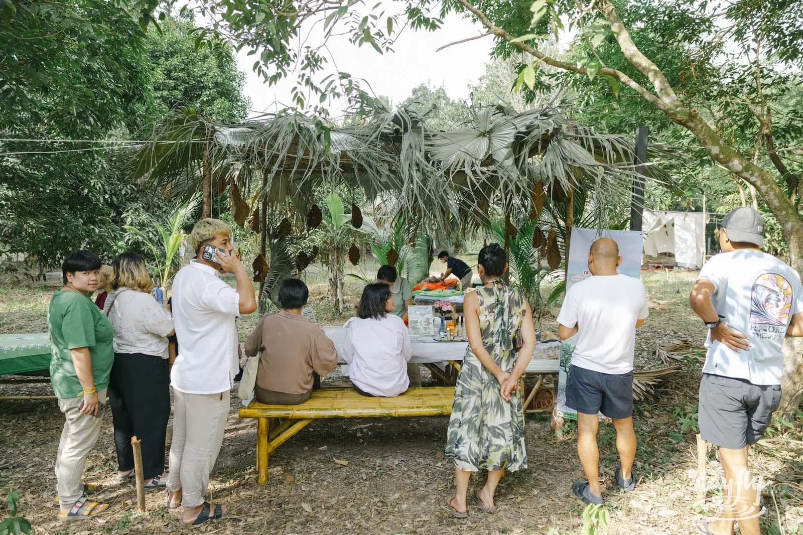 Visitors stand and sit beside a bamboo table beneath a leafy shelter.