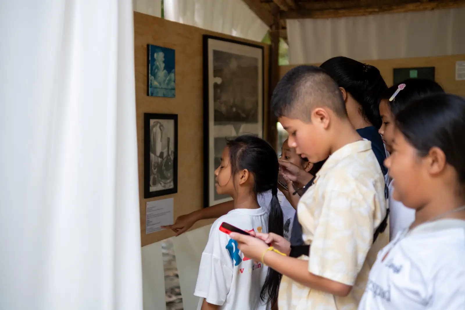 Students in white shirts stand close to framed drawings and prints hung on a cloth wall, reading the labels.