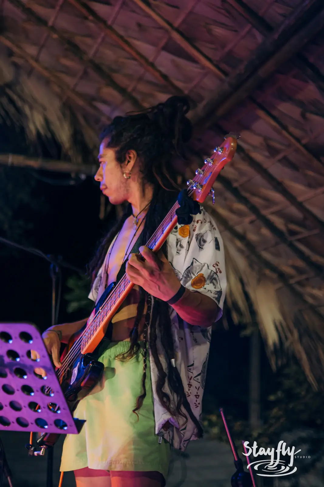 Bassist with dreadlocks playing under thatched roof.