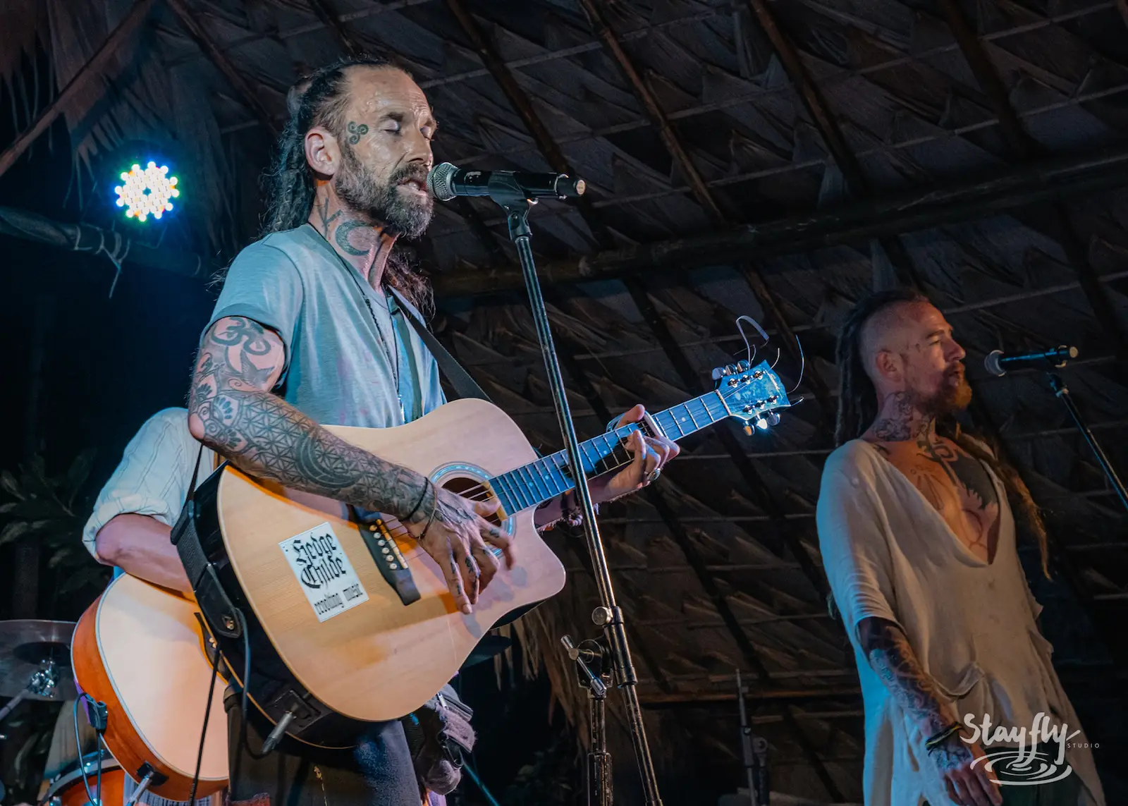 Man singing with acoustic guitar under thatched roof.