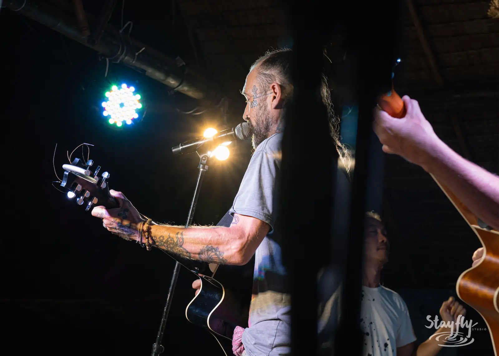 Tattooed guitarist singing under thatched stage roof.