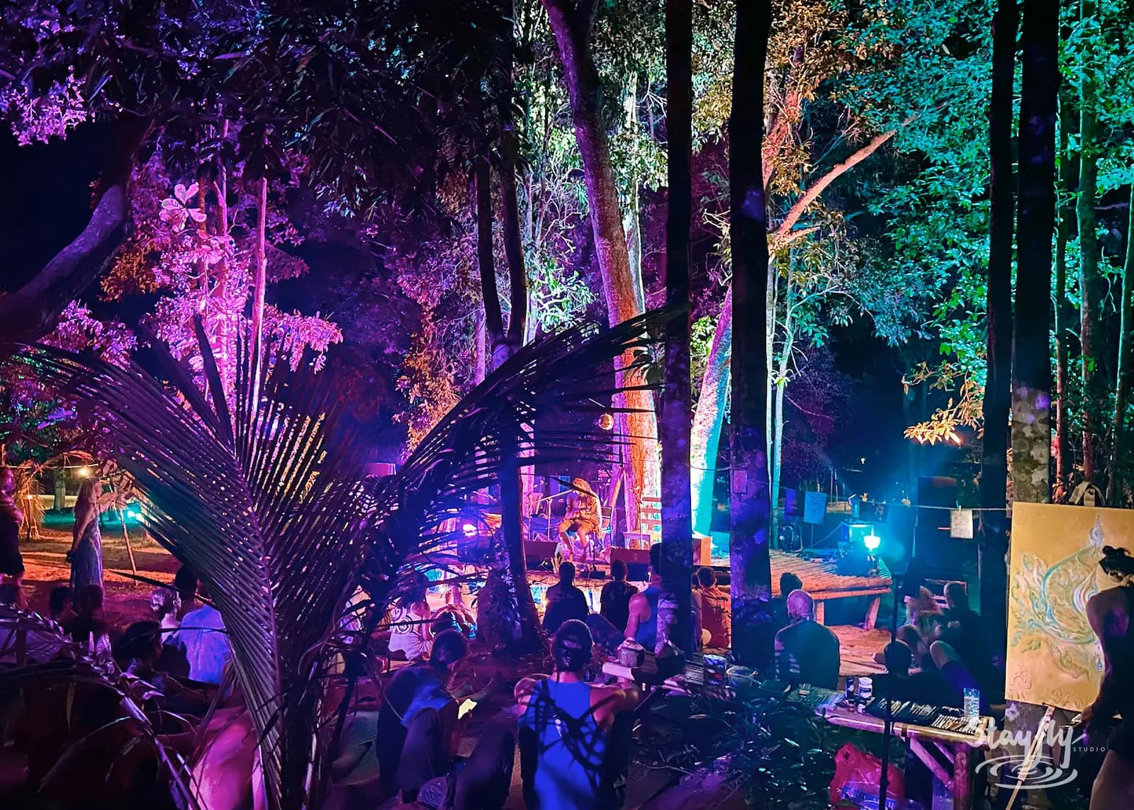 Crowd gathers under purple-lit trees at night.