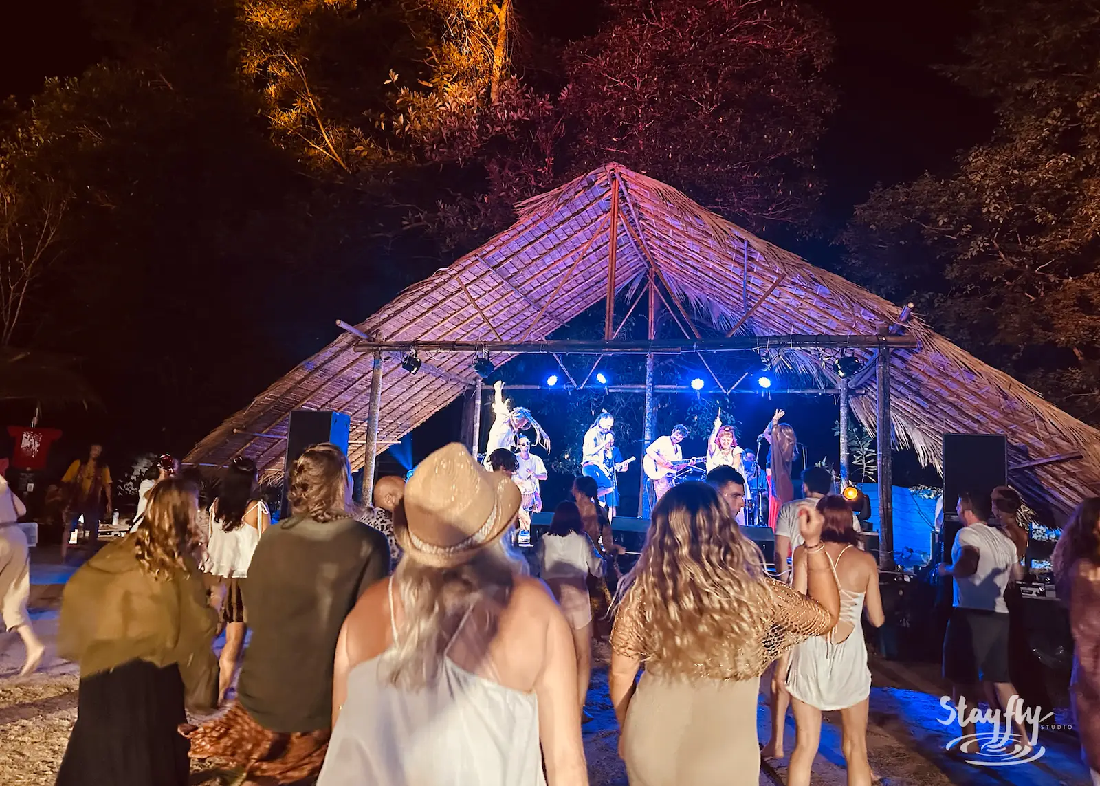 Band plays bamboo stage, crowd dances at night.