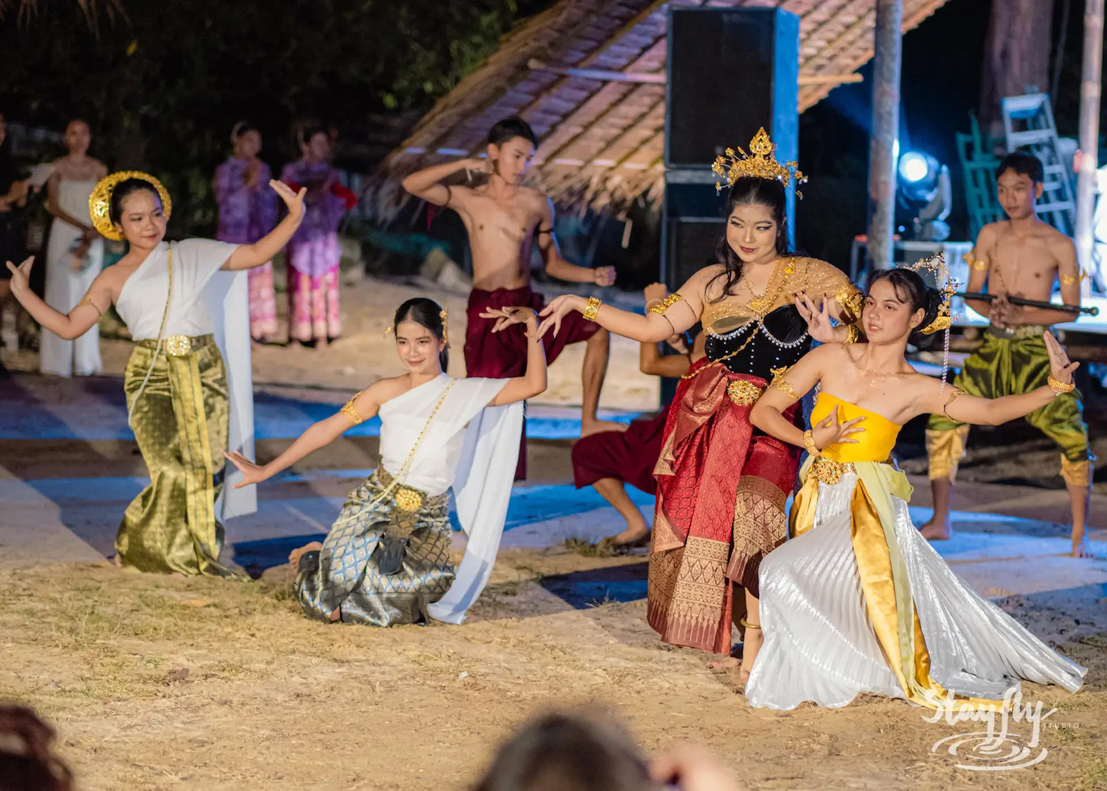 Dancers in Thai classical costume perform on the sand at night beside a thatched hut.