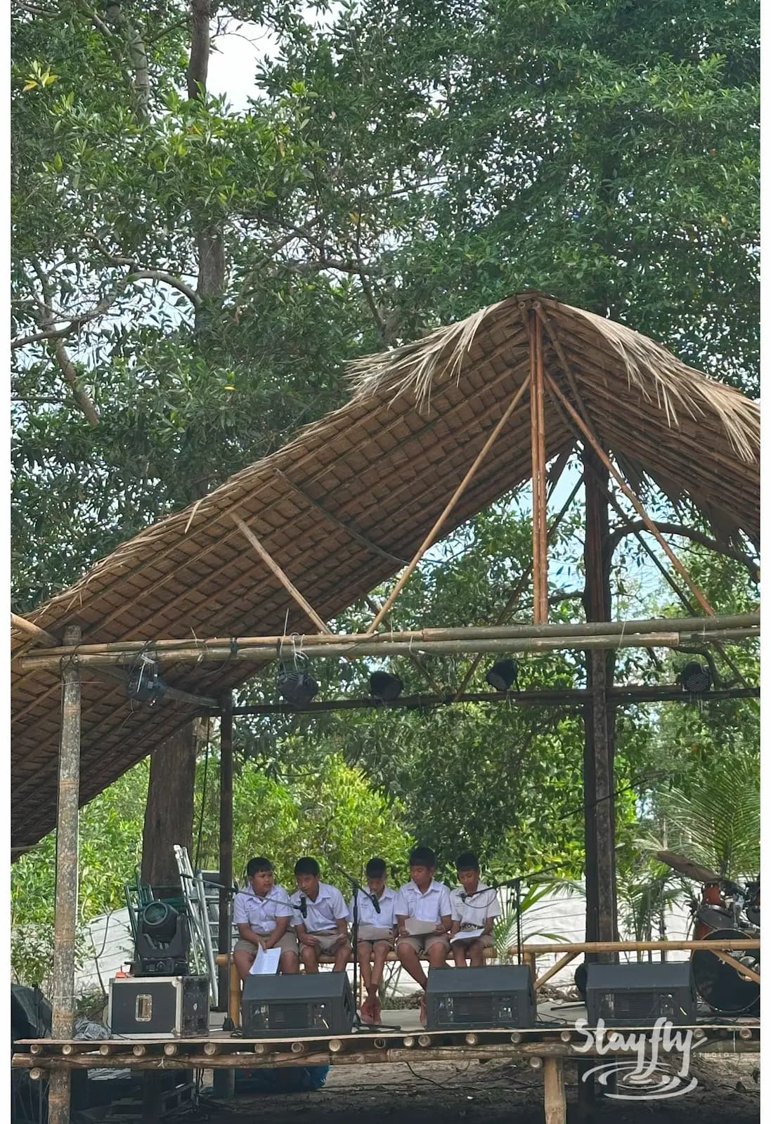 Students seated on outdoor stage under thatched roof.