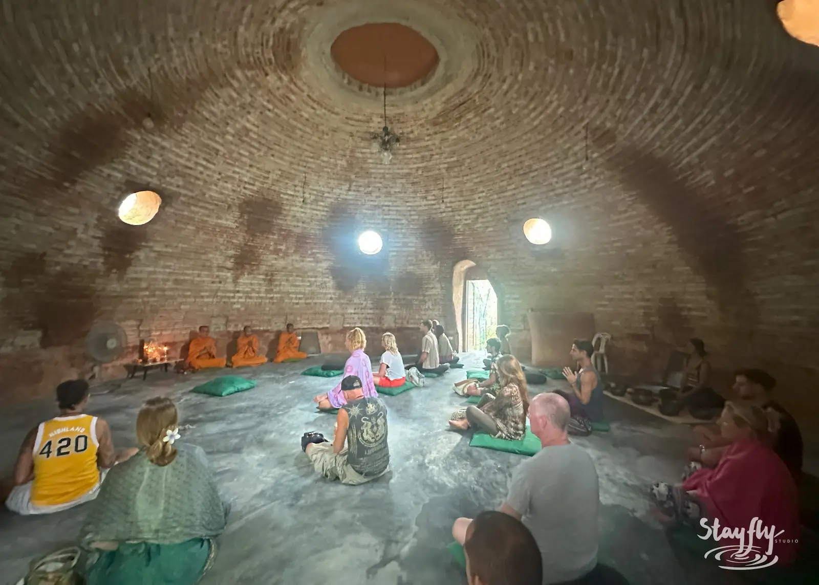 Three monks in orange robes sit against the wall of the brick Dome as people gather on cushions around them.