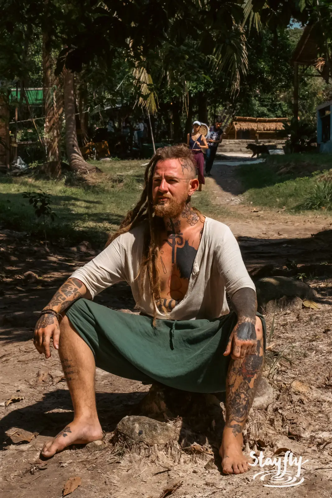 Tattooed man sits on tree stump outdoors.