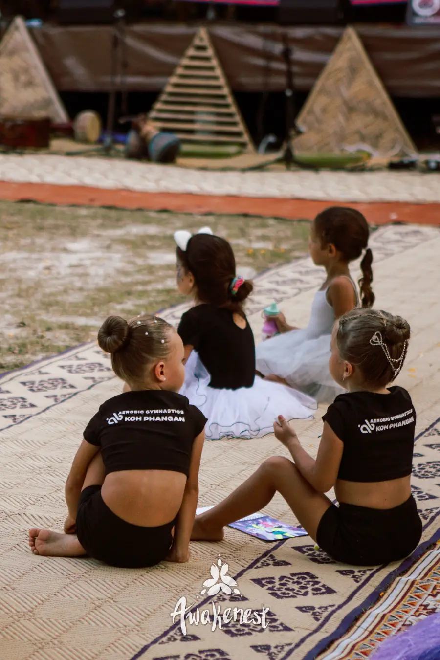 Girls in gymnastics club shirts seated on mat.
