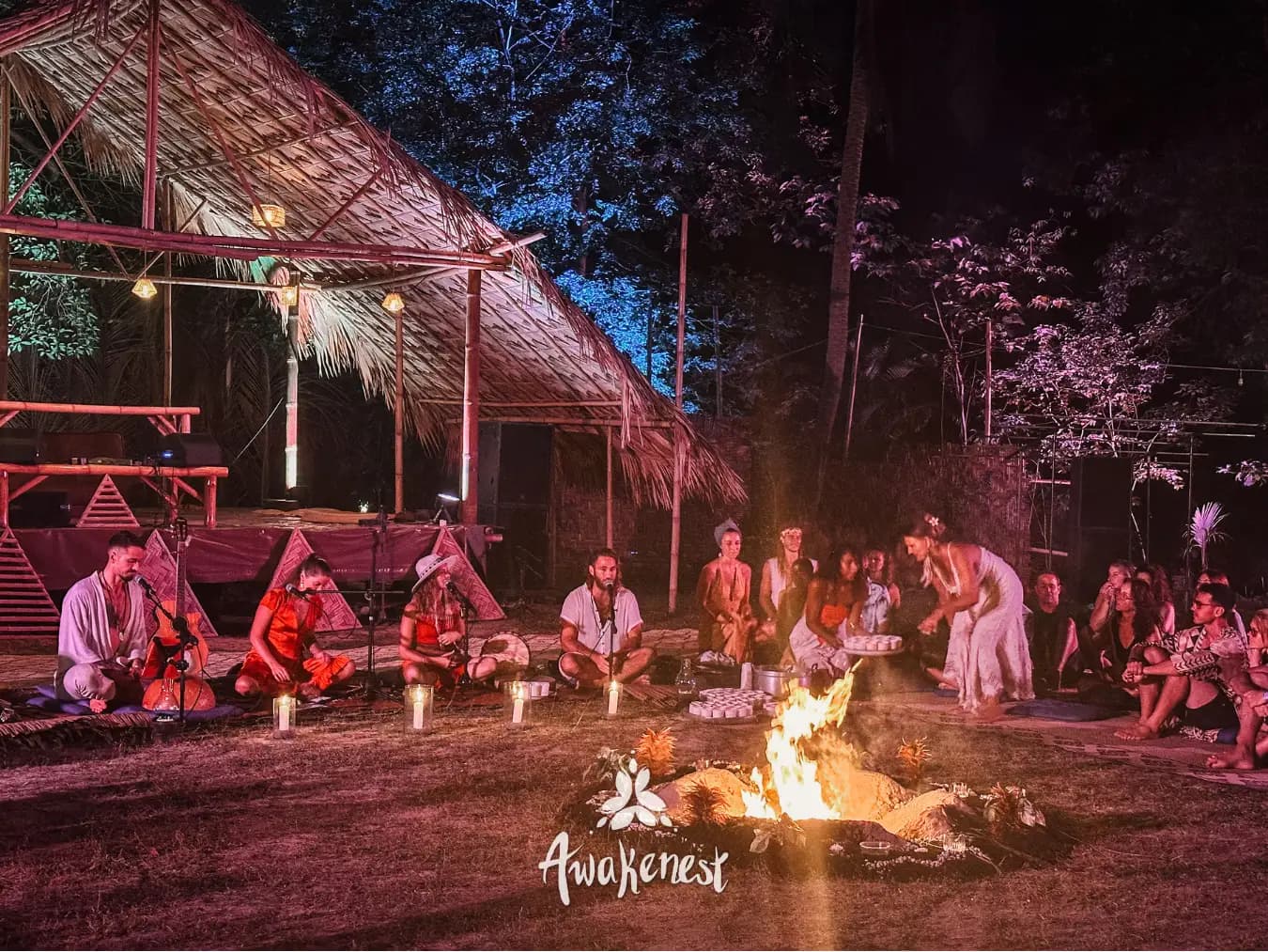 Robed musicians sit and play on the ground beside a fire at night.