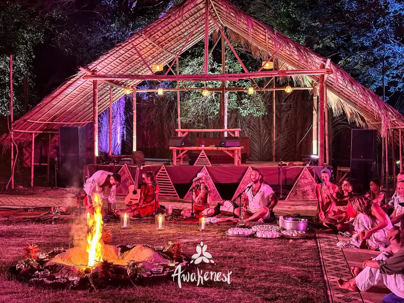 A fire burns on open ground in front of a seated circle beneath an open thatched pavilion at night.