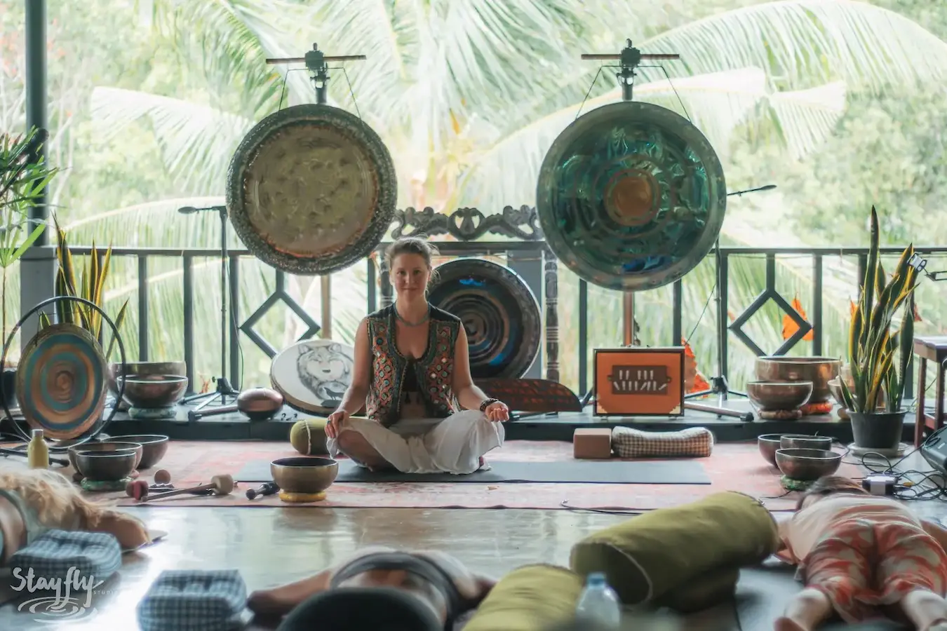 A practitioner in white sits between two gongs on a rug with hands at the heart.