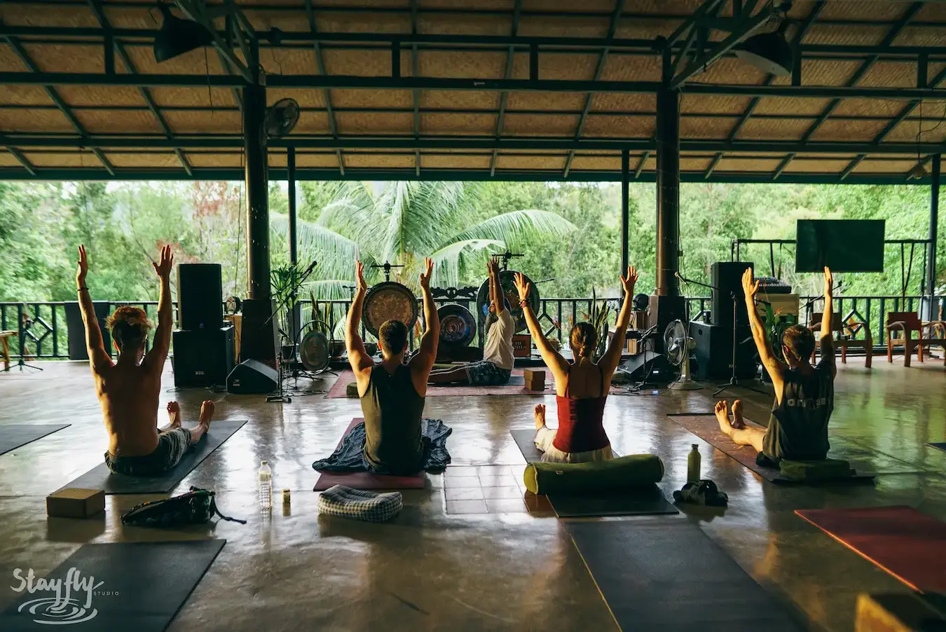 A yoga class raises its arms together in the open pavilion with gongs hung behind.