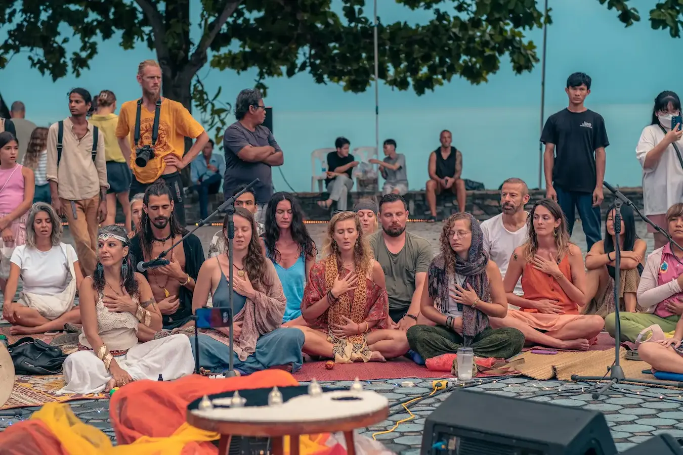 People sit together on mats by the water with their hands at their hearts.