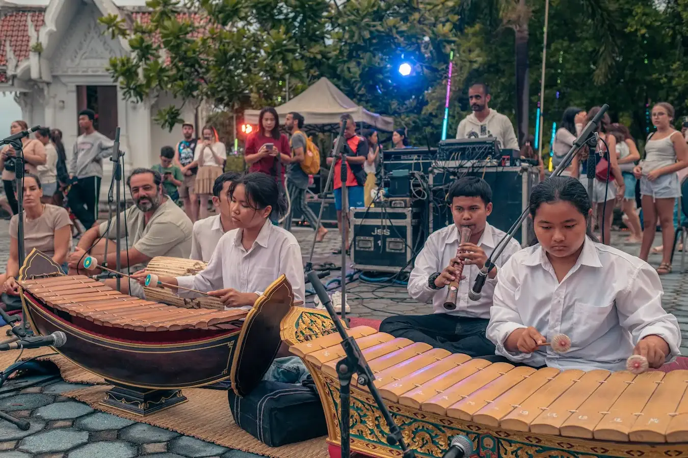 Young musicians in white play two Thai xylophones and a flute on a mat at the pier while people gather behind them.