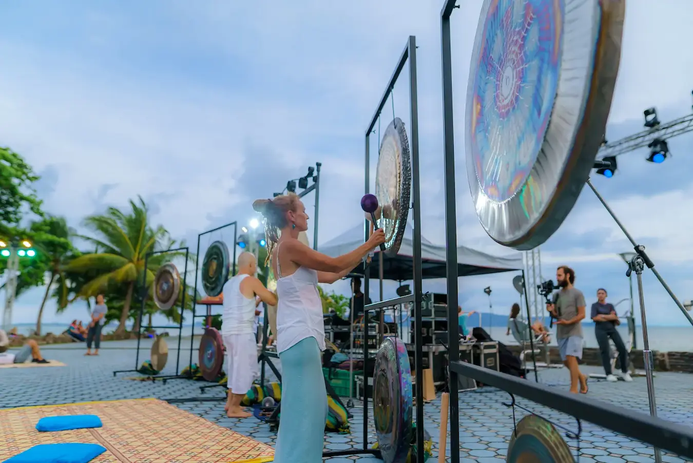 Large painted gongs stand in a row beside the sea, one of them being played.