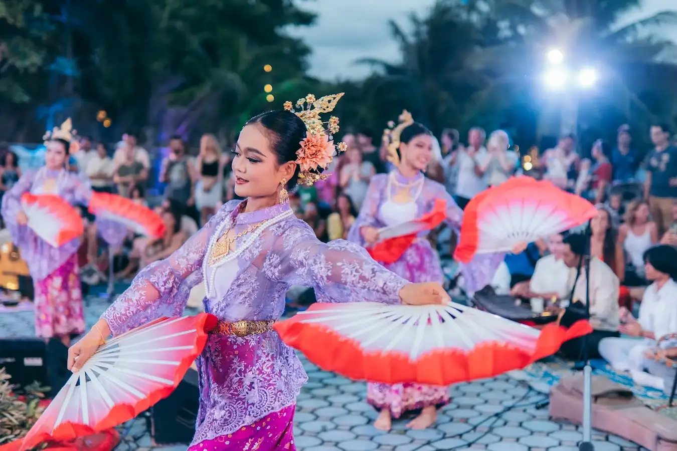Dancers with red and white fans perform in front of a seated gathering at dusk.