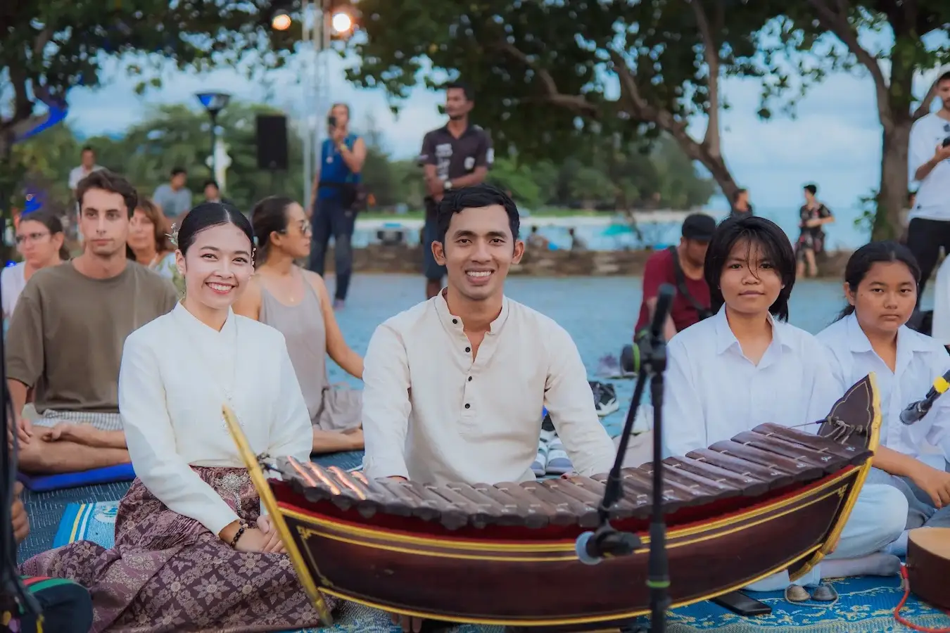 Young musicians in white play a boat-shaped Thai xylophone seated beside the sea.
