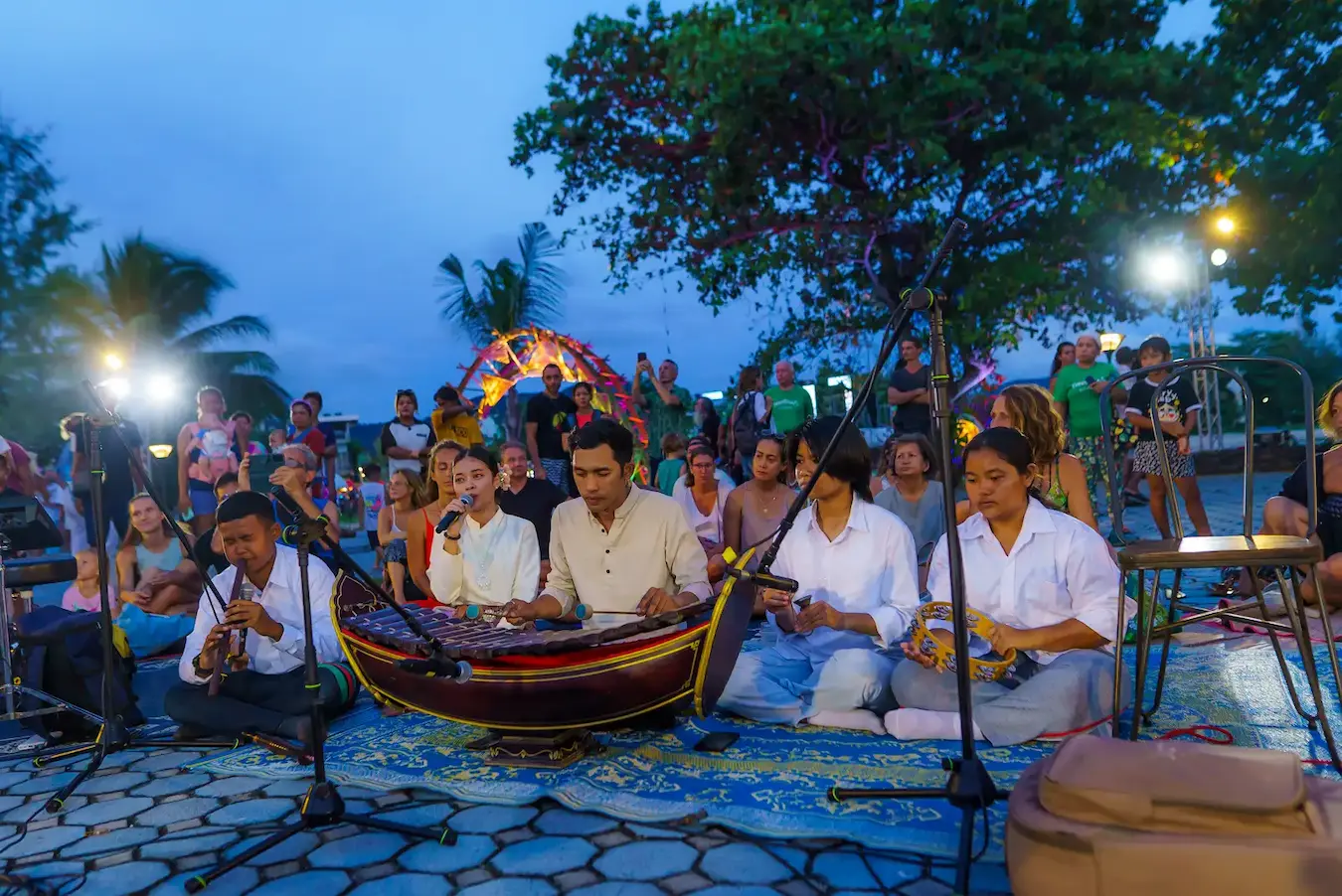 Musicians play a Thai xylophone and flute on a mat at dusk while a singer holds the microphone and the crowd listens behind them.