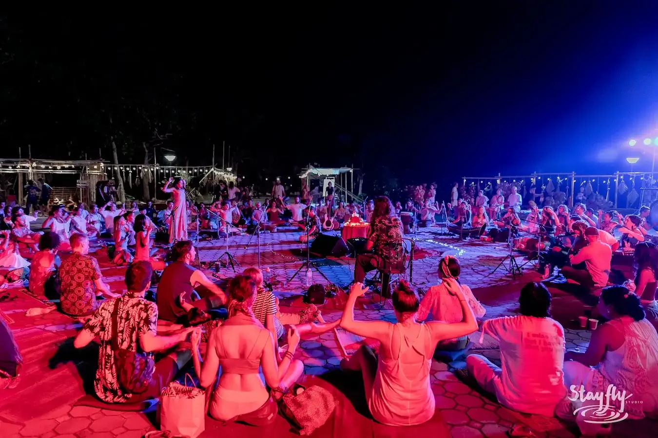 A wide night gathering seated on mats at the pier under pink and blue light.
