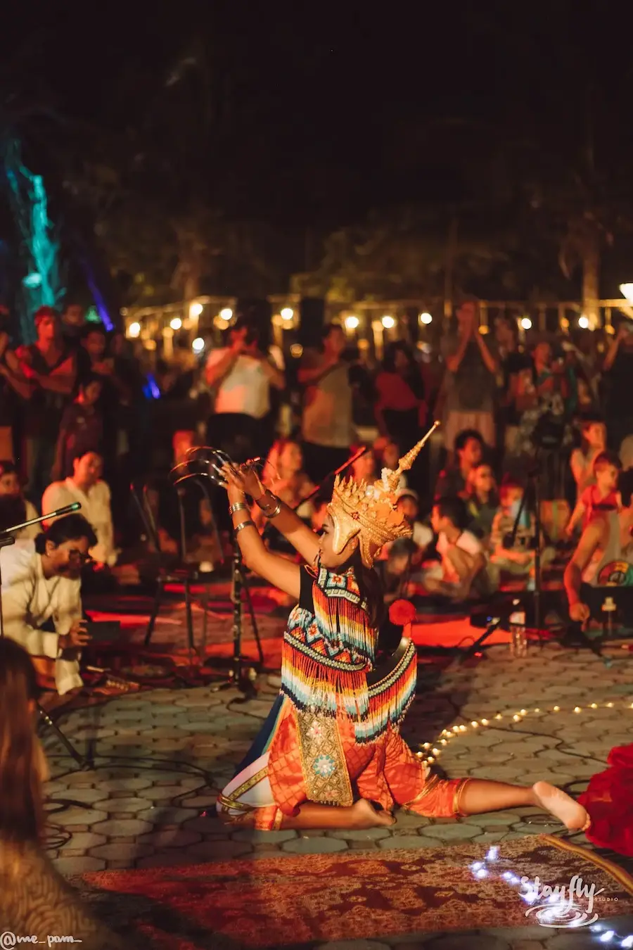 A dancer in an ornate crown performs inside a seated gathering under strings of lights at night.