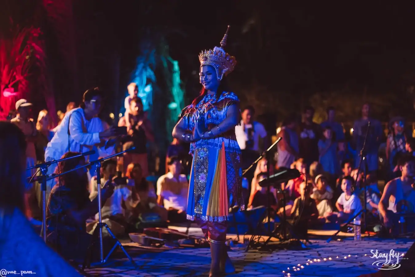 A Nora dancer in a beaded costume and tall crown performs among the audience under coloured light at night.