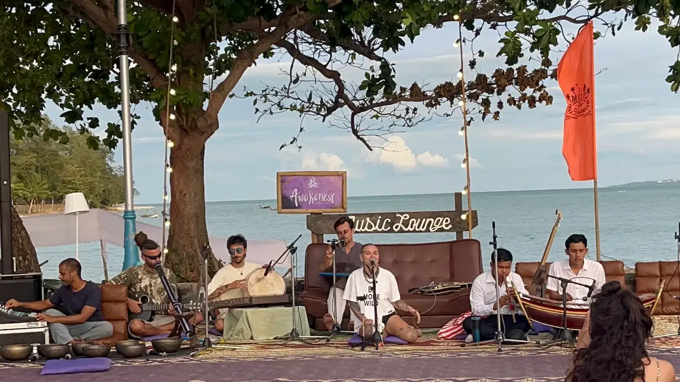 Young musicians in white play a Thai flute and xylophone beside guitarists, singing bowls and a singer under a tree by the sea.