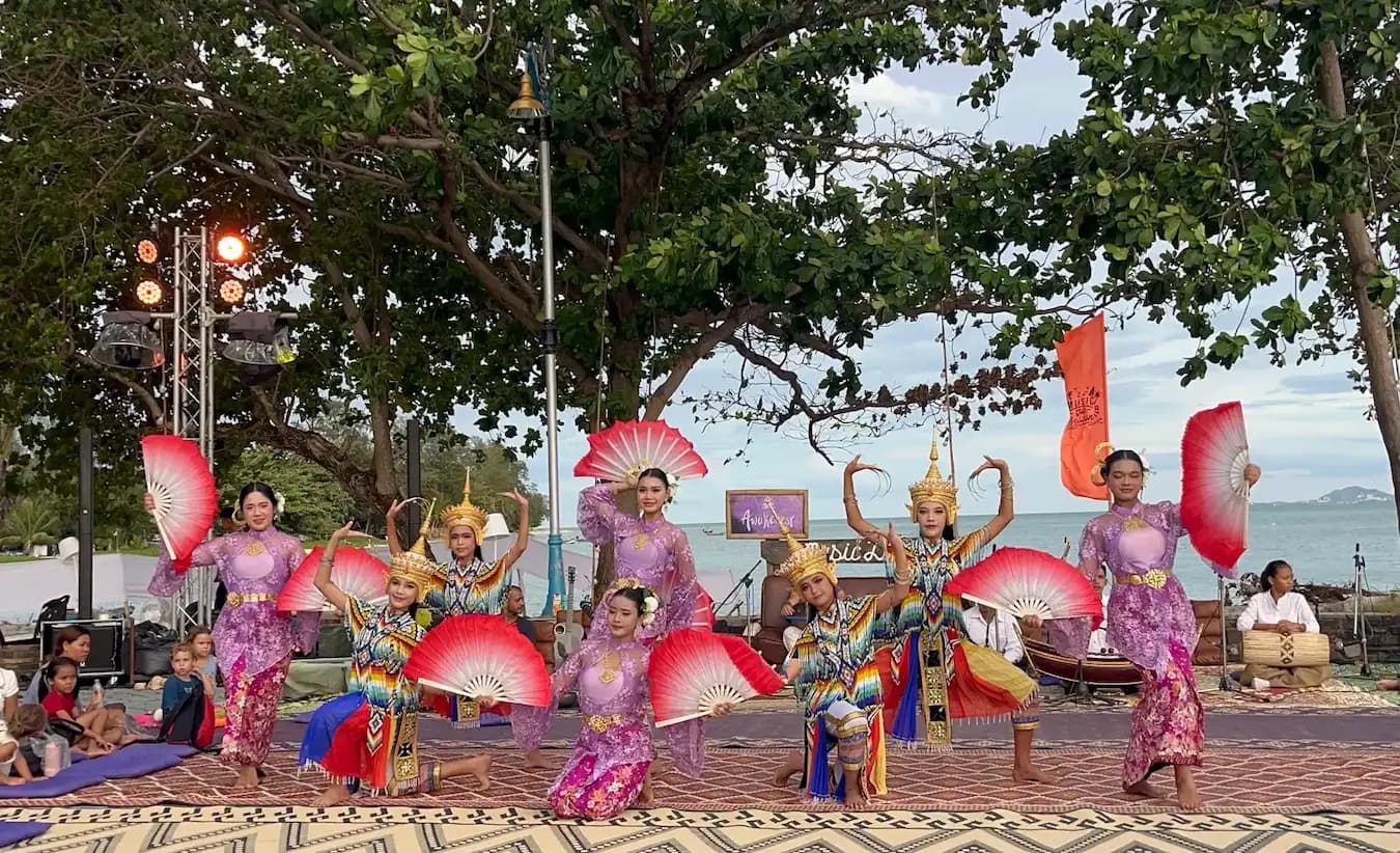 Dancers in purple costumes raise red and white fans beneath a tree by the sea.