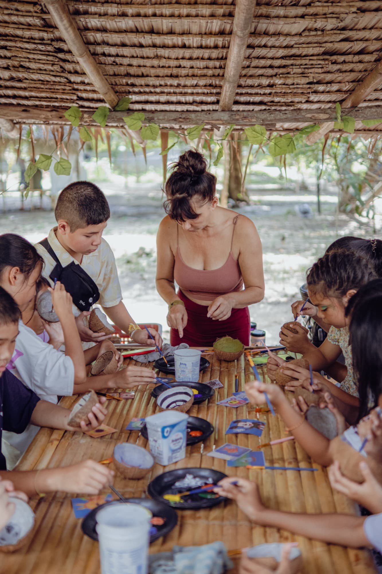 Children paint coconut shell bowls around a long bamboo table under a thatched roof while a facilitator leans in to help.
